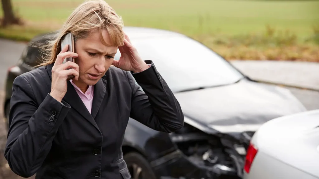 A woman looks distressed while holding her head, standing next to a car with visible damage after an accident, and speaking on the phone