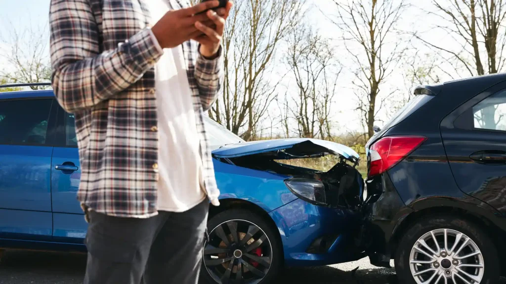 Person on phone standing near a blue car with rear-end damage from an accident.